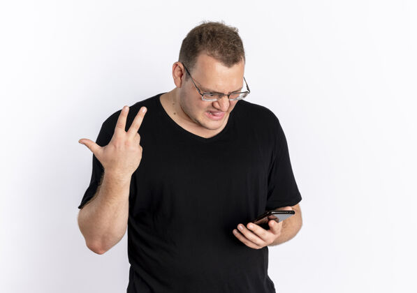 手机一个戴着眼镜 穿着黑色t恤的超重男人看着他的智能手机屏幕 困惑和不快的手站在白色的墙上手困惑眼镜