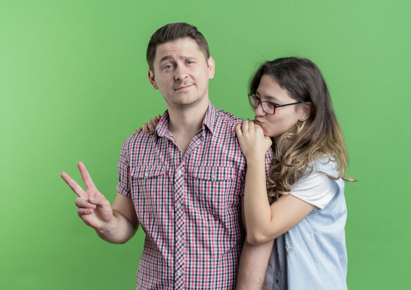 站立一对穿着休闲服的年轻夫妇站在一起 一个男人在绿色的墙上展示v形标志Vsign年轻快乐