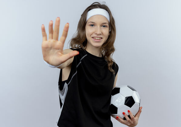 年轻身穿黑色运动服的年轻健身女孩 头上系着足球 站在白色的墙上微笑着张开手做停车标志立场停止使