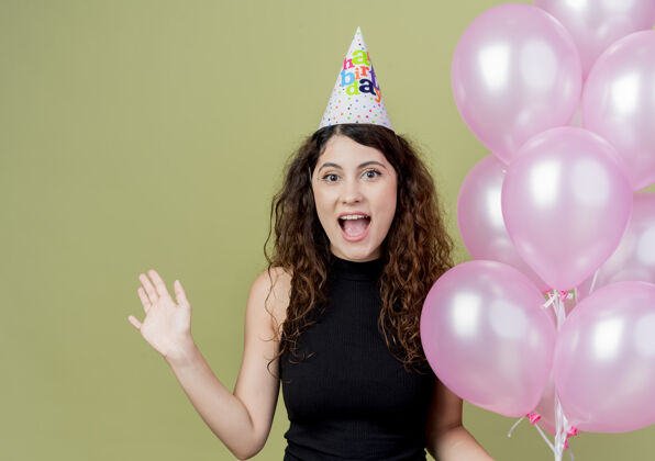 举行一位年轻漂亮的女士 卷发 戴着节日帽 手持气球 举手 高兴而兴奋地微笑着 生日派对的概念站在光墙上手空气提高