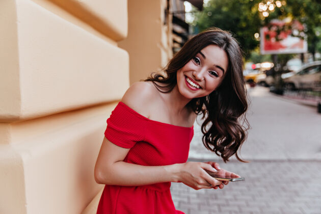表情一个迷人的女人 卷发 站在大楼附近 手里拿着电话一个黑发 穿着红裙子的女孩 对着镜头笑着肖像城市放松