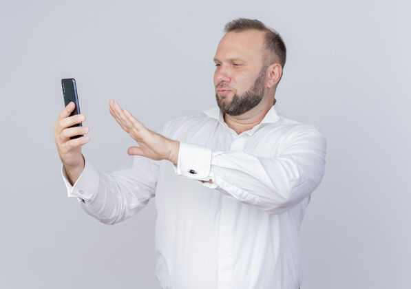 握着一个留着胡子的男人穿着白衬衫拿着智能手机打视频电话看着屏幕做防御手势站在白墙上视频防御男人