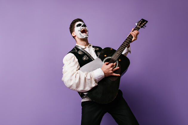 情感面部艺术的家伙想象自己的摇滚音乐家和情感姿态与吉他室内肖像的人在紫丁香墙上西班牙墨西哥幽灵