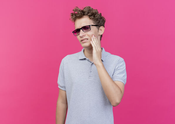 马球一个戴着黑眼镜 穿着灰色马球衫的年轻人站在粉红色的墙上 看着不舒服 摸着脸颊 牙疼看脸颊眼镜