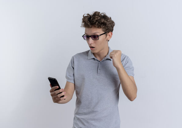 马球穿着灰色马球衫的年轻人看着智能手机屏幕 握紧拳头 站在白色的墙上 快乐而兴奋屏幕看拳头