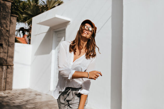 情感穿着灰色裤子和超大号衬衫的积极时尚的女人在白色建筑和棕榈树旁的街道上玩得很开心棕榈树帽子温暖