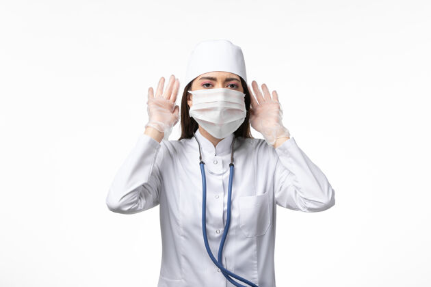 听力正面图女医生穿着白色无菌医疗服 带着口罩 因冠状病毒试图听到白墙上的疾病冠状病毒-大流行性病毒病大流行面罩成人