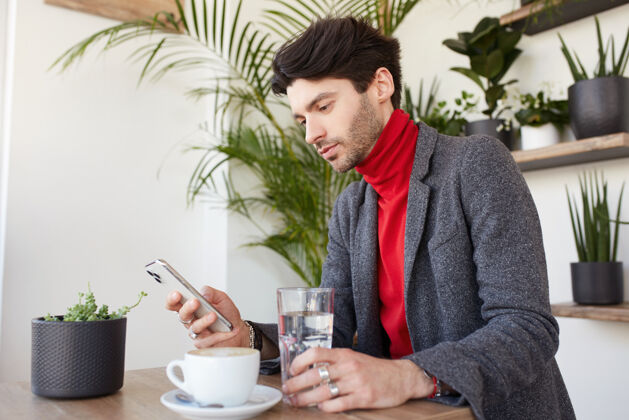 胡须年轻迷人的棕色头发的男人坐在城市咖啡馆手机夹克优雅