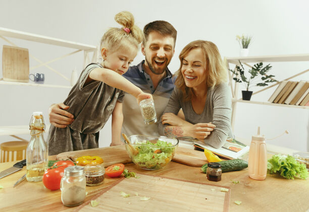 吃饭可爱的小女孩和她美丽的父母在家的厨房里切蔬菜 一边微笑着做沙拉积极童年蔬菜