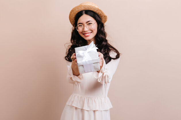 工作室拍摄开朗的韩国女士展示礼物戴着帽子的亚洲模特拿着礼物盒米色背景高兴微笑