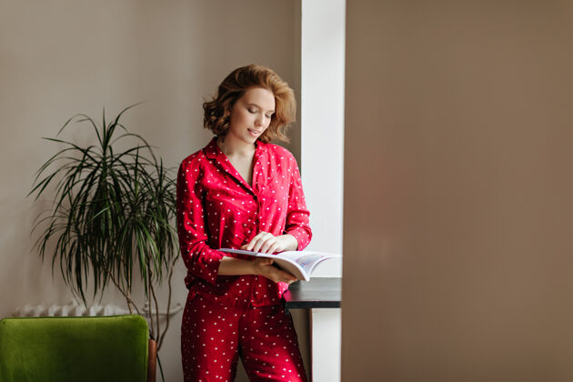 扶手椅早上看杂志的美丽沉思的女人穿着红色睡衣的漂亮女人的室内镜头杂志公寓寒冷