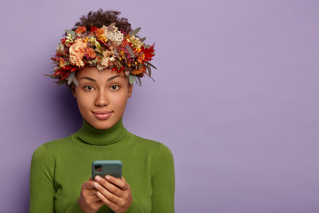 使用你好秋天可爱的女人头上戴着秋天的花环 穿着绿色休闲服 用手机花环黑色电话