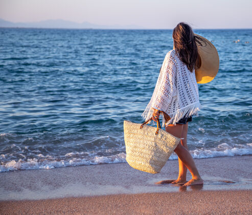 海岸一个手里拿着帽子和柳条袋的女孩走在海边暑假的概念日落散步女人