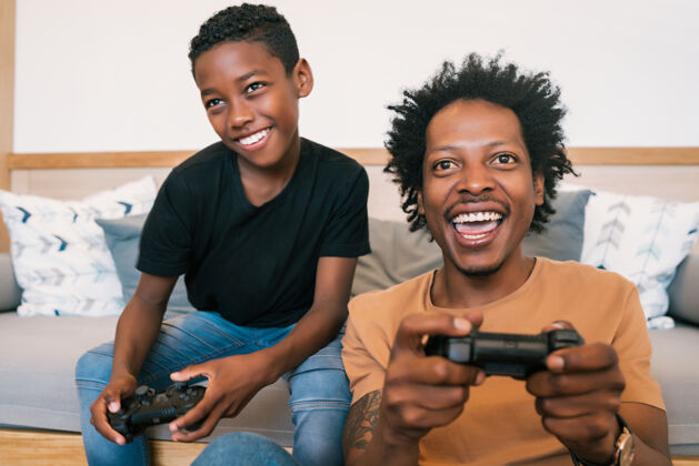 游戏快乐的非洲裔美国人的父亲和儿子坐在沙发上 在家里一起玩游戏机家庭和技术概念美国非洲裔美国人科技