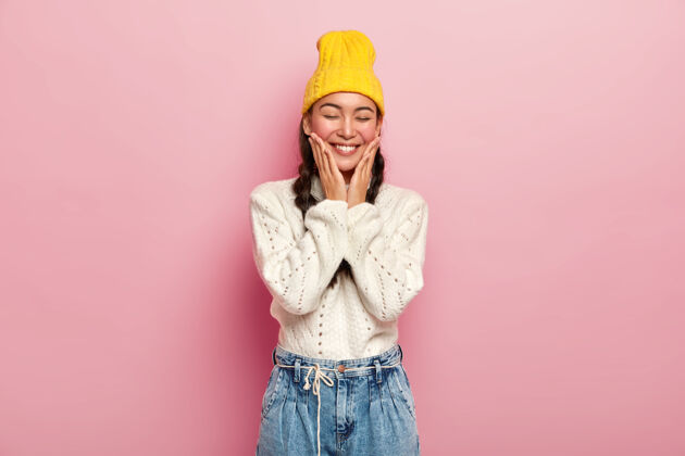 女人迷人的韩国年轻女子摸着脸颊 有着满意的表情 闭着眼睛 害羞 戴着黄色的帽子和针织毛衣套头衫一件女孩
