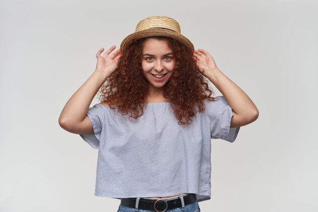 青少年有魅力的成年红头发女孩的肖像 卷发穿着条纹露肩上衣和帽子触摸她的帽子和微笑隔着白色的墙壁孤立微笑皮肤20多岁