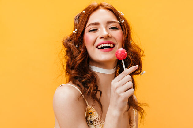 姜红色卷发的女人笑着吃着棒棒糖橙色背景上的年轻女人的画像野花美丽化妆品