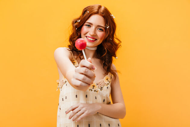 黄色背景身着黄色太阳裙的快乐女人手持粉色糖果橙色背景上头发上插着鲜花的女人肖像时尚花时尚