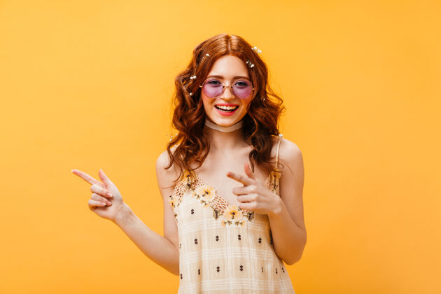 魅力戴太阳镜的红发女子将手指指向左边身穿黄色上衣的女子在孤立的橙色背景下微笑着性感野花美丽