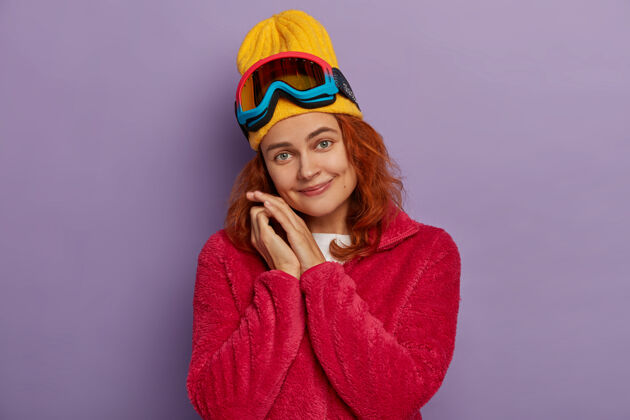 紫色美女歪着头 戴着黄色帽子和红色夹克 玩得很开心 用滑雪眼镜高兴地看着隔离在紫罗兰墙上的相机酒窝年轻冬天
