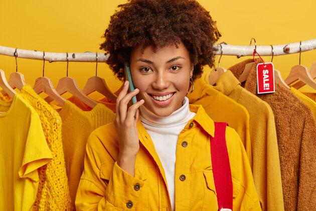 商店可爱的卷发女人的特写镜头打电话 笑容满面 穿着黄色夹克欢呼客户举行