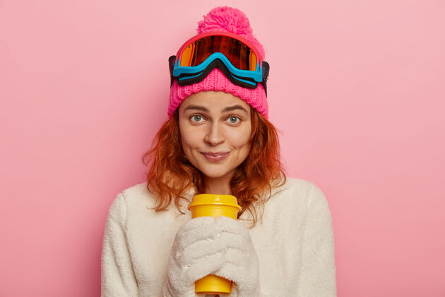 酒窝女孩滑雪穿温暖的冬季服装 白色手套 举行外卖咖啡 粉红色背景隔离女士帽子红发