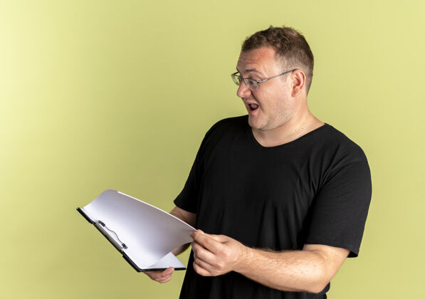人一个戴着眼镜的胖男人 穿着黑色t恤 剪贴板上有空白页 站在浅色的墙上 满脸幸福地看着他们超重页光
