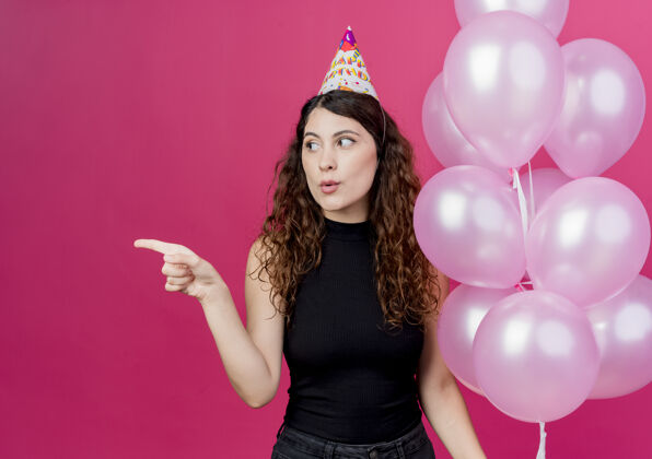 气球一位年轻漂亮的女士 头上戴着节日帽 手里拿着一堆气球 手指着站在粉色墙上的生日派对概念女人年轻壁板