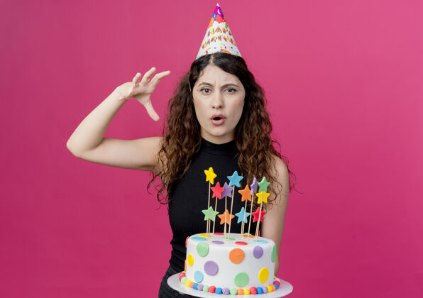 蛋糕年轻漂亮的女人卷发戴着节日帽拿着生日蛋糕站在粉红色的墙上不高兴的生日派对概念年轻不高兴看着