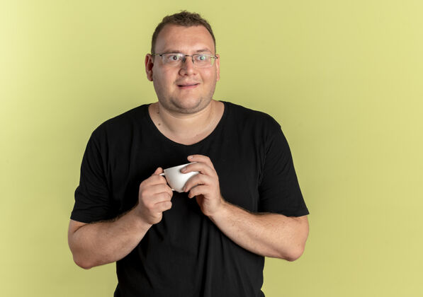 咖啡一个戴眼镜的胖男人 穿着黑色t恤 手里拿着咖啡杯 一边看一边情绪激动 一边开心地站在轻质的墙上超重眼镜男人