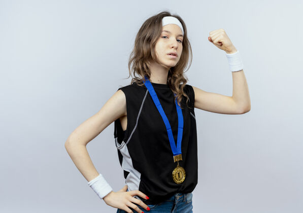 看身穿黑色运动服的年轻健身女孩 头上戴着头带 脖子上戴着金牌 举起拳头 站在白色的墙上 看上去像个胜利者 自信满满健康周围拳头