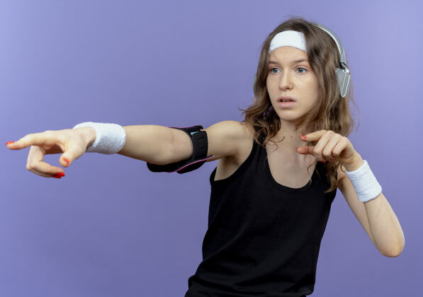 目录身穿黑色运动服的年轻健身女孩 戴着头带和智能手机臂带 食指指向蓝色墙壁上方的一侧运动服移动头带