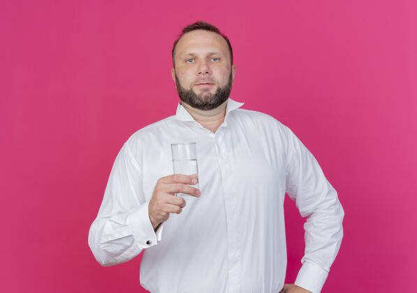 脸满脸胡须 身穿白衬衫 端着一杯水的男子站在粉红色的墙上 表情严肃看着戴着站着