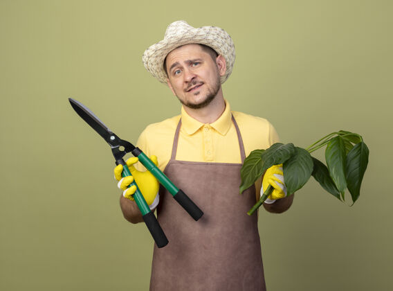 植物年轻的园丁 穿着连体衣 戴着帽子 戴着工作手套 手里拿着植物和树篱剪 满脸笑容手套微笑剪子