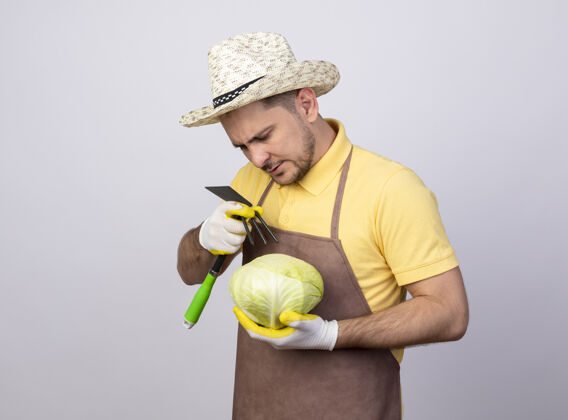 严肃年轻的园丁 穿着连体衣 戴着帽子 戴着工作手套 手里拿着卷心菜和床垫 表情严肃男人手套花园