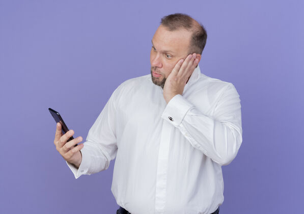 严肃一个留着胡子的男人 穿着白衬衫 拿着智能手机 站在蓝色的墙上看着屏幕 严肃的脸被弄糊涂了屏幕移动抱着