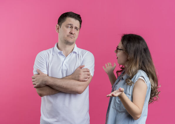 交叉一对穿着休闲服的年轻夫妇双手交叉放在胸前 他的女朋友站在粉红色的墙上和他争吵休闲女朋友争论