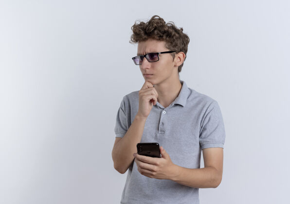 拿着戴着黑眼镜 身穿灰色马球衫的年轻人手持智能手机 手放在下巴上 神情沉思地站在白墙上站着移动的放在一边