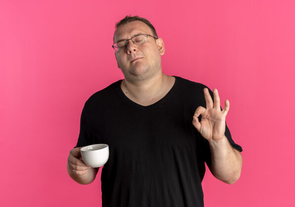 咖啡一个戴眼镜 身穿黑色t恤 手持咖啡杯的超重男子站在粉红色的墙上高兴地打着“一切正常”的手势拿着眼镜杯子