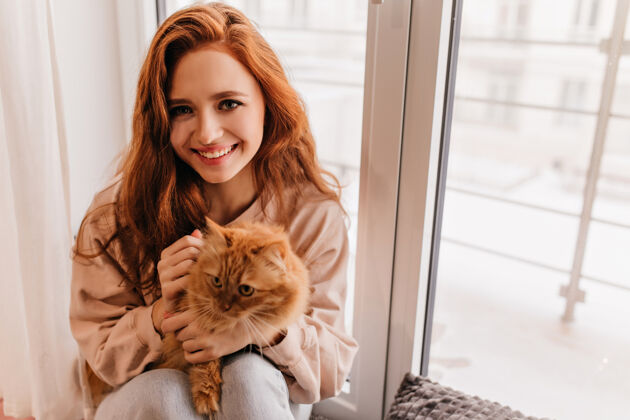 积极美丽的姜女微笑着站在窗边室内镜头中 迷人的女孩抱着一只红色的毛茸茸的猫姜玩耍年轻女子