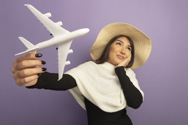 向上戴着白色围巾戴着夏帽的年轻女子展示玩具飞机抬头思考积极夏天紫色玩具