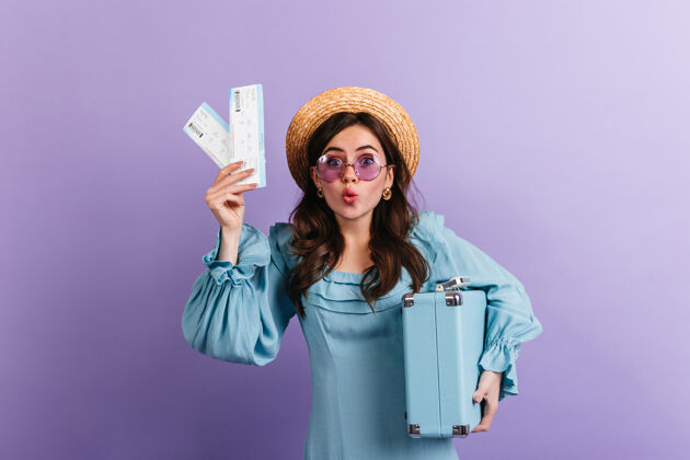 自信一个戴着紫丁香眼镜的滑稽女人惊奇地盯着她 展示她的机票和蓝色复古手提箱可爱肖像时尚