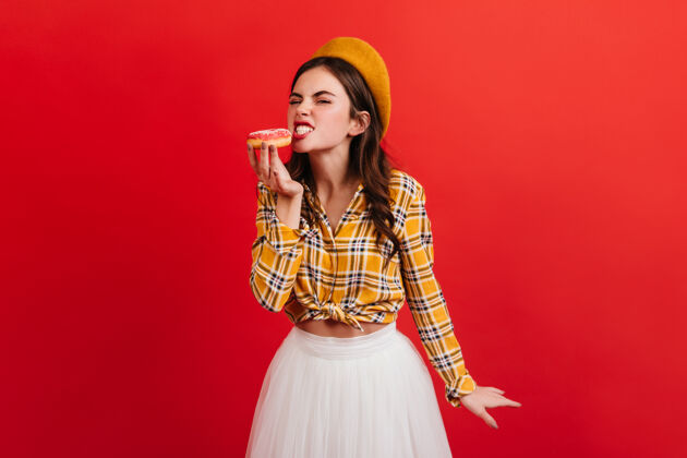 模特穿着时髦服装的饥肠辘辘的女孩咬着草莓甜甜圈明亮的墙上挂着一幅身穿格子衬衫 头戴橙色贝雷帽的女人的画像积极服装肖像