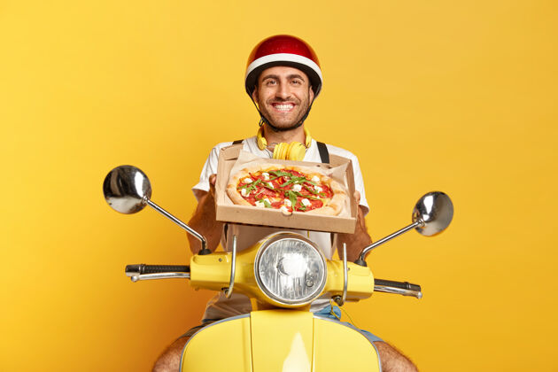 送货满意的送货员带着头盔驾驶黄色踏板车 手里拿着比萨饼盒举行纸板姿势