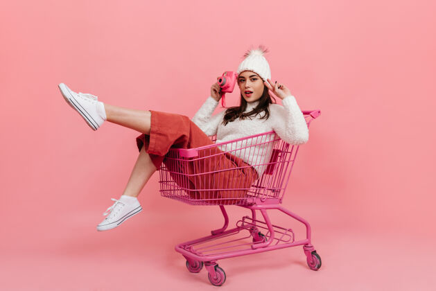 成人一个穿着针织衣服的年轻女孩坐在超市的手推车里 对着粉色的相机摆姿势波浪发模特时尚