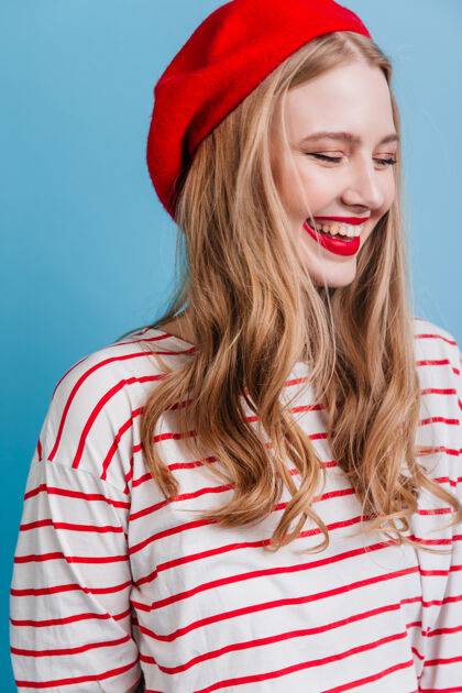 游戏穿着贝雷帽和条纹衬衫的情绪化女孩在蓝色的墙上欢笑幸福的金发女模特发型法国时尚