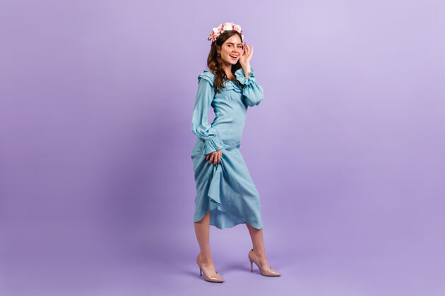 蓝色连衣裙可爱的微笑着的女士穿着缎蓝色的衣服女人在淡紫色的墙上抚摸她的脸玫瑰脸卷发