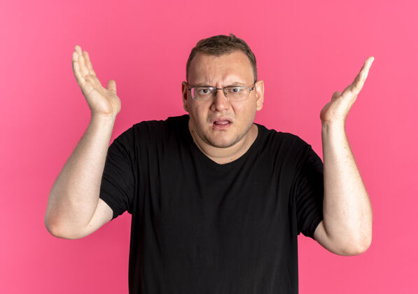 手臂一个戴眼镜 身穿黑色t恤的超重男子站在粉色的墙上 困惑地 不悦地举起双臂询问或争论争论问站