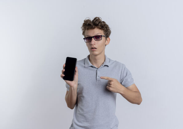 展示一个戴着黑眼镜 穿着灰色马球衫的年轻人站在白墙上 用手指着智能手机 困惑地看着它马球眼镜年轻