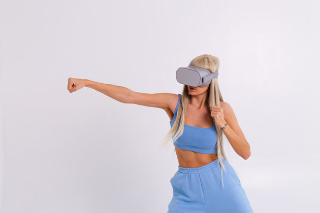 设备摄影棚照片中 一个年轻漂亮的女子穿着温暖的蓝色时尚西装 戴着虚拟现实眼镜 在白色舞台上打拳击玩家科技搏击